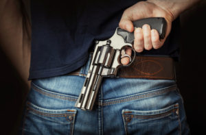A person in a blue shirt and jeans stands against a dark backdrop, silver revolver at their waist, finger cautiously off the trigger—an image often encountered by gun crime defense attorneys as they unravel the stories behind such perilous moments.