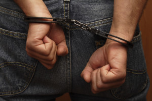 A person wearing jeans is shown from behind with their hands in fists, handcuffed and awaiting the expertise of a marijuana defense attorney. The background is out of focus.