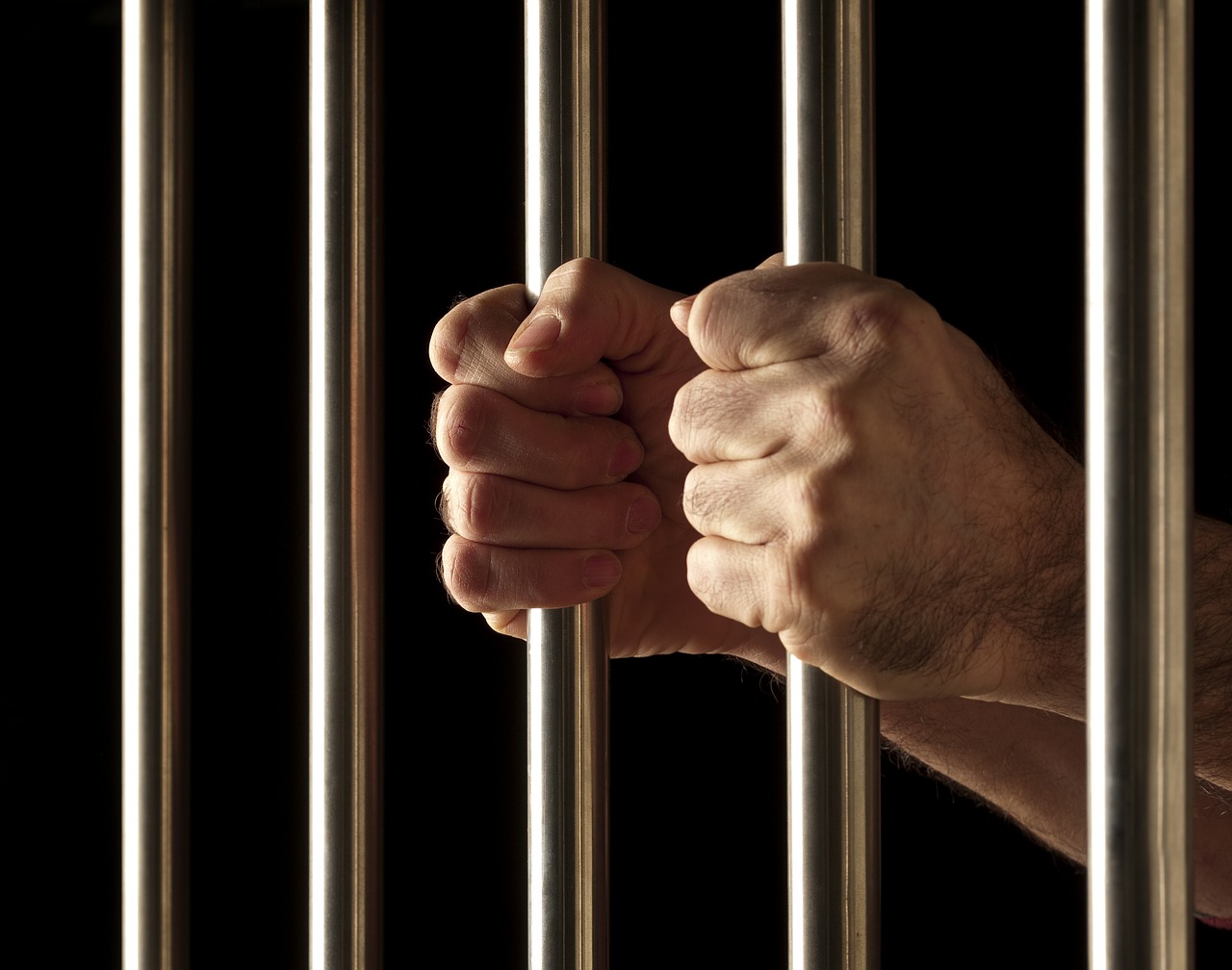 Two hands gripping metal prison bars against a dark background symbolize confinement and imprisonment, highlighting the importance of bail as a means of securing temporary freedom. The image conveys a sense of captivity and restraint.