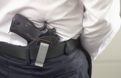 A person is wearing a white shirt and dark pants with a black belt. A handgun, subject to NY SAFE Act regulations, is tucked into the back of their waistband, partially visible.
