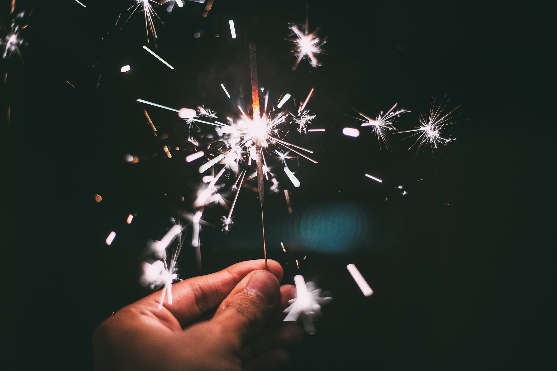 Close-up of a hand holding a lit sparkler against a dark background. Bright sparks radiate outward, reminiscent of the fireworks often enjoyed on festive nights, though some celebrations must heed New York State's laws where certain fireworks are illegal.