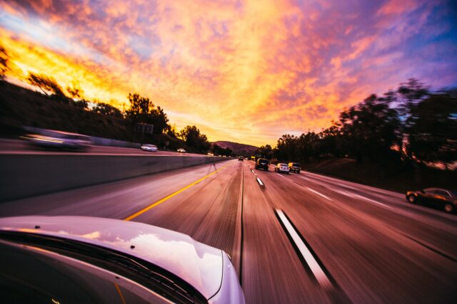 As the car speeds down a multi-lane highway at sunset, vibrant orange and purple clouds fill the New York State sky. The road, lined with scattered trees and other vehicles, creates a sense of motion, reminding drivers to heed traffic violations penalties in their journey.