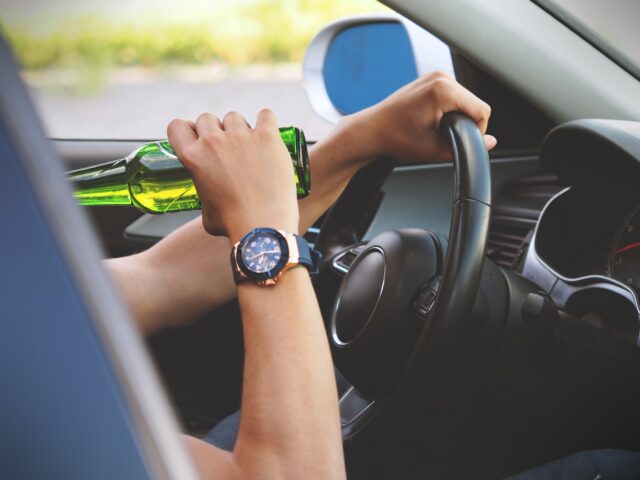 In New York State, DWI penalties are strict; even a first offense can lead to severe consequences. The image shows a person holding a green bottle while driving, their watch visible on the wrist as they grip the steering wheel.