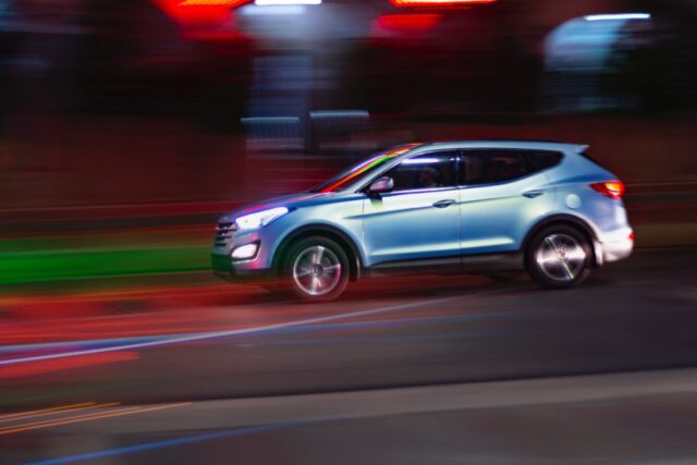 A silver SUV is driving swiftly through an urban area at night, as law enforcement vigilantly patrols the streets. The background blurs with streaks of red and green light, highlighting the vehicle's motion and capturing the vibrant essence of urban nightlife.