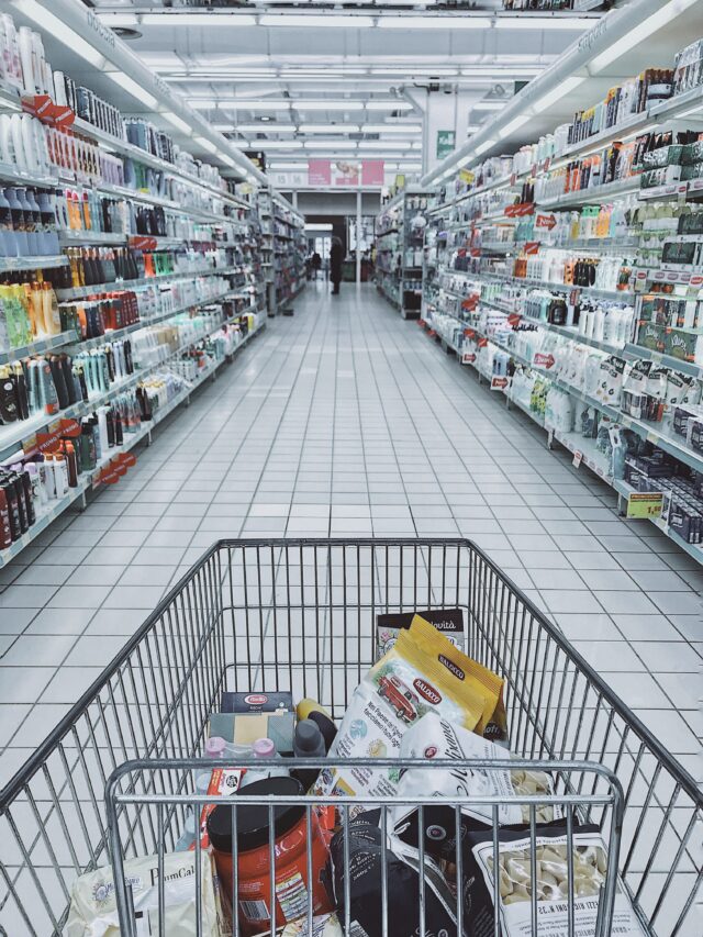 A view from a shopping cart filled with various groceries under the bright fluorescent lighting of a New York State supermarket aisle. Shelves are stocked with products, like bottles and boxed goods, yet any thoughts of larceny charges remind shoppers to leave without facing penalties.