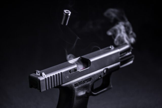 A semi-automatic handgun with smoke rising from the barrel is shown against a dark background. In a scene reminiscent of Nassau County's struggle against gang violence, a bullet casing is ejected mid-air, suggesting recent firing.