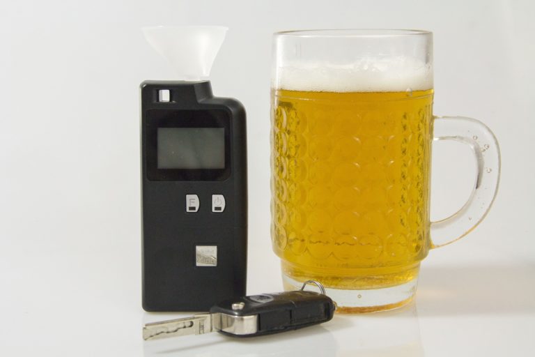 A breathalyzer for BAC testing, a key fob, and a frothy beer mug rest on a white surface. The breathalyzer features a digital display and mouthpiece, highlighting its importance in New York DWI scenarios.