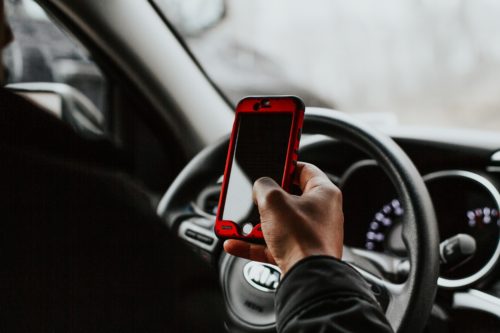 A person holding a smartphone with a red case while driving showcases a potential cellphone violation. The car's dashboard and steering wheel are visible, with the hand on the phone as the blurred background highlights the vehicle's interior—a reminder to consult a cell phone violation lawyer if needed.