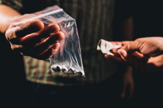 In the dimly lit background, a hand quietly passes a small plastic bag filled with white pills, while another reaches out with folded money. The shadowy transaction hints at the need for a methamphetamine possession attorney.
