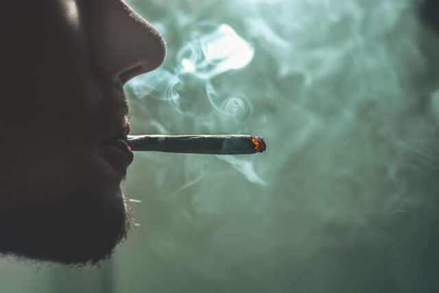 Close-up of a person’s face in profile, with a lit joint in their mouth. Smoke gently swirls around their mouth and nose against a blurred greenish background, reminiscent of the hazy atmosphere often recounted by a Marijuana Defense Attorney.