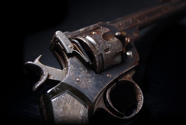 Close-up of an old, rusted revolver with visible corrosion and wear, evoking thoughts of gun crime. The focus is on the trigger and barrel area, highlighting the texture and age of the metal. The dark background emphasizes its weathered appearance, reminiscent of cases handled by defense attorneys.