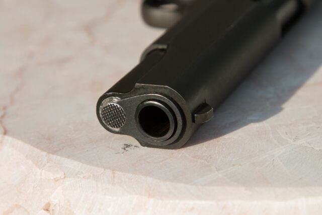 Close-up of a black handgun muzzle on a light-colored, textured surface. The focus is on the gun's barrel and front sight, with intricate details visible in the metal finish, highlighting its role in personal defense for those with a gun permit. Nassau County Gun Permit Defense concept image
