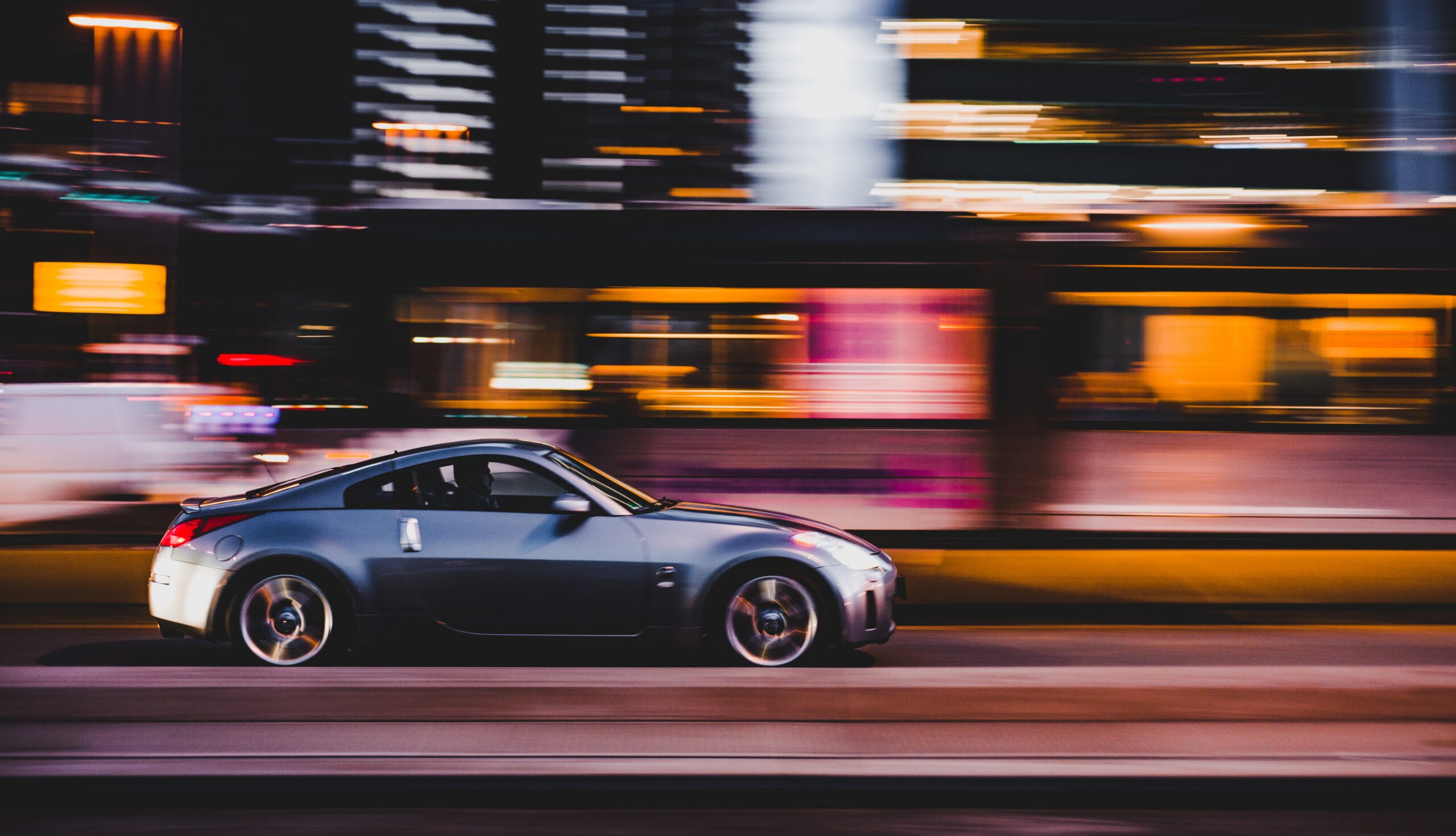 A sleek silver sports car speeds down a city street at night, with blurred lights and buildings in the background, suggesting motion and vibrant urban energy. The driver might soon need a speeding ticket lawyer to keep up with their nocturnal adventures.