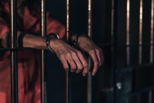 In the dimly lit cell, a person in an orange prison uniform stands behind bars with hands in handcuffs, their arms extending through the cold metal. The somber atmosphere underscores the gravity of their situation—one that urgently calls for an experienced violent crime lawyer.
