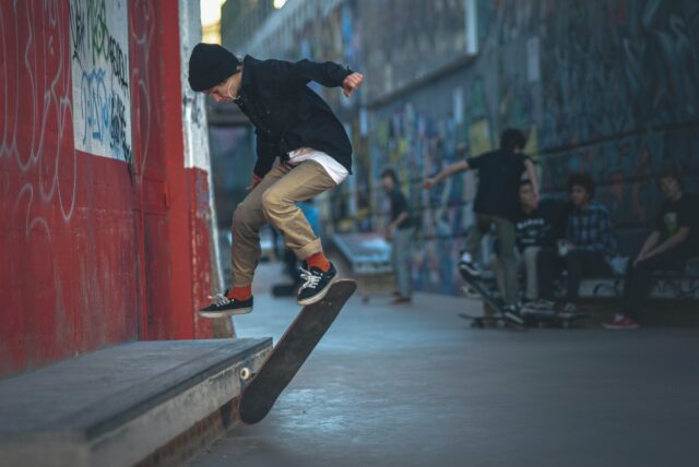 A skateboarder in a black hoodie and beige pants performs a trick on a ledge in an urban skate park, surrounded by graffiti-covered walls. A group of people sit on a bench watching, their chatter momentarily distracting from tales of juvenile crimes that color the city’s streets.