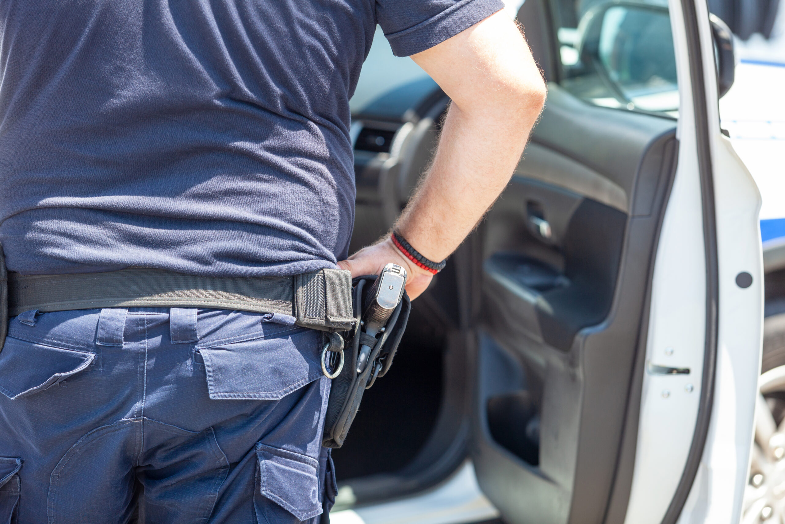 A law enforcement officer in a navy uniform reaches for a handgun in their holster, standing beside an open car door. The focus is on the officer's hand and equipment belt, underscoring New York Vehicle Search procedures and legal rights during such searches.