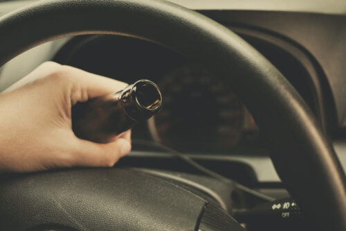 A hand grips a brown glass bottle inside a car, resting on the steering wheel, while the dimly lit interior suggests it’s nighttime. A sense of relief hangs in the air, like the calm after having a DUI dismissed.