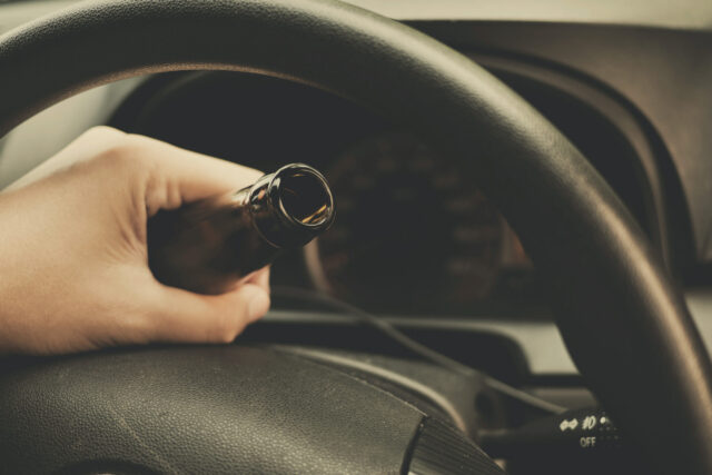 A hand grips a brown glass bottle inside a car, resting on the steering wheel, while the dimly lit interior suggests it’s nighttime. A sense of relief hangs in the air, like the calm after having a DUI dismissed.