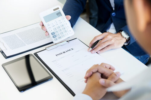 An insurance agent and client sit across from each other at a desk. The agent holds a calculator displaying "499" and discusses DWI car insurance options, pointing to an insurance contract on a clipboard. A tablet and documents are also on the desk.