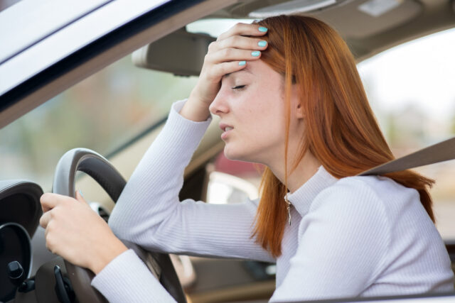 A woman with red hair, possibly under 21, sits in the driver's seat of a car, wearing a seatbelt. She seems stressed or tired, resting her forehead on her hand with eyes closed—perhaps pondering a DWI risk as the steering wheel looms ahead.