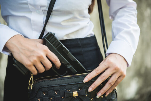 A person in a white shirt and dark skirt discreetly prepares to carry a firearm, placing a black handgun into a black purse with gold accents. Only the torso and hands are visible.