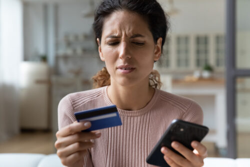 A woman with a concerned expression holds a credit card in one hand and a smartphone in the other, sitting indoors, possibly worried about credit card fraud penalties.