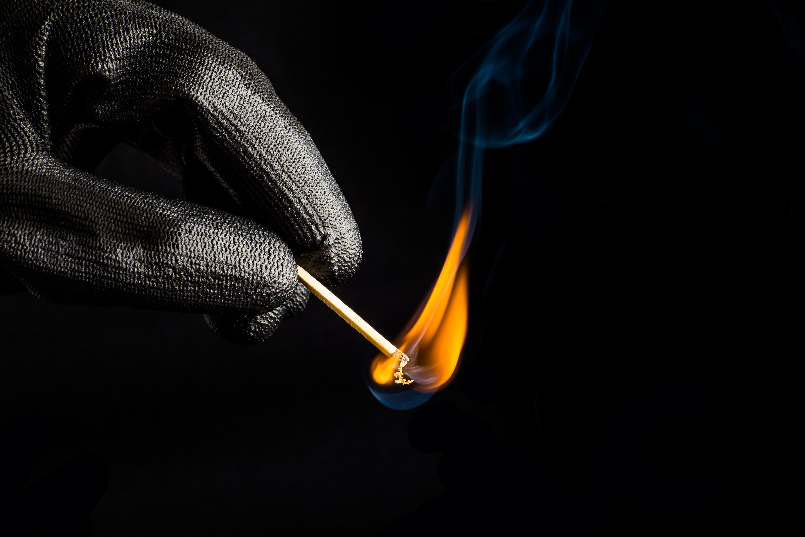 A gloved hand holds a lit matchstick, producing a small orange flame and wisps of blue smoke against a dark background—a stark reminder of the severe arson penalties for unlawful fire-setting.