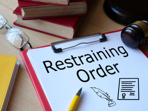 A clipboard with a document labeled "Restraining Order" sits on a desk alongside a gavel, books, reading glasses, and legal-themed icons—a scene symbolizing the process of obtaining a restraining order.