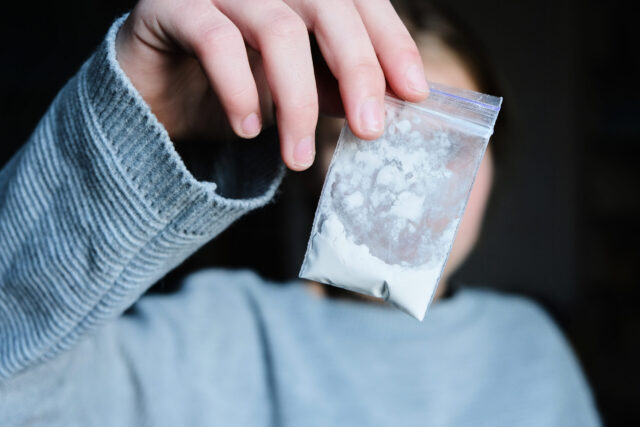 A person in a gray sweater holds up a small clear plastic bag containing a white powdery substance, with their face out of focus—a scene often considered when discussing defenses to drug possession charges.