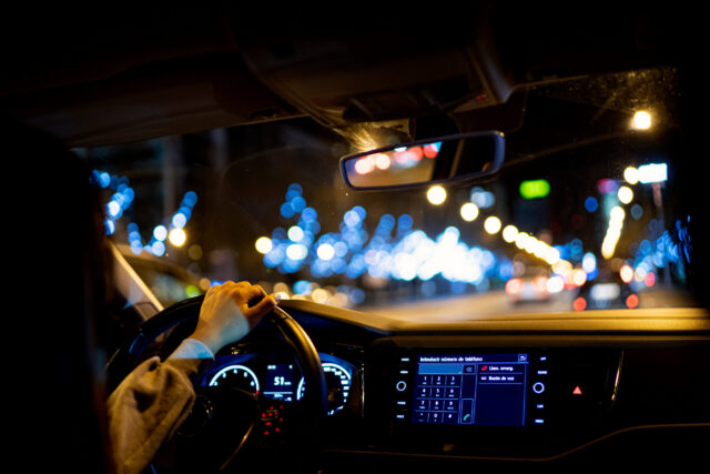 A person drives a car at night through a city street, their left hand on the steering wheel. The illuminated dashboard contrasts with blurred lights outside—a reminder to always obey New York traffic laws and avoid hit-and-run accidents in New York.