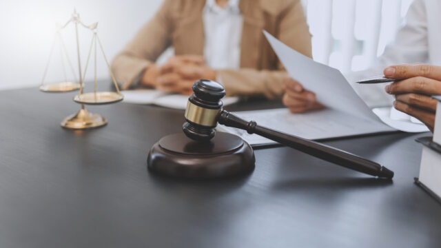 A wooden judge's gavel and sound block on a table, with a scale of justice and two people in the background reviewing documents related to domestic violence charges in New York.