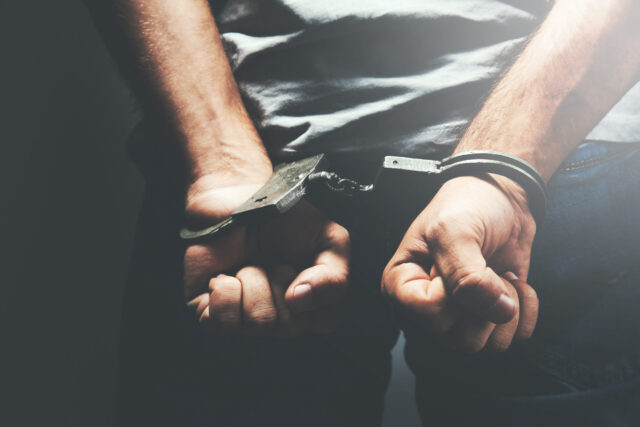 Close-up of a person’s hands in handcuffs behind their back, arrested for possession of stolen property, wearing a gray shirt and jeans, with a dark background and light shining from above.
