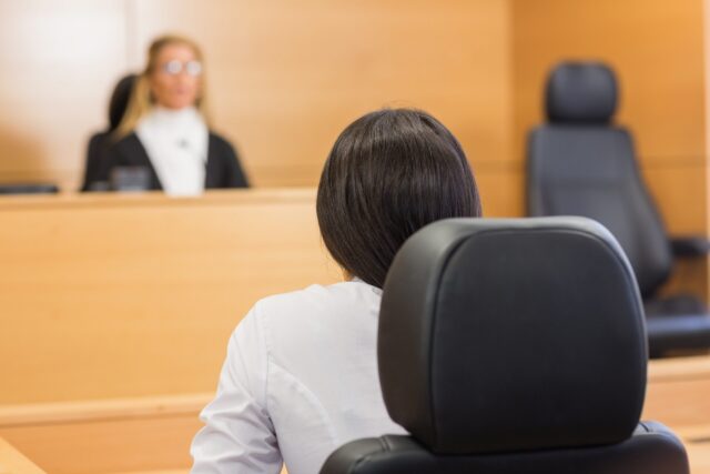 A person with dark hair sits in a courtroom, facing a judge at the bench, suggesting a legal proceeding or criminal trial and highlighting the potential risks of representing yourself in court.