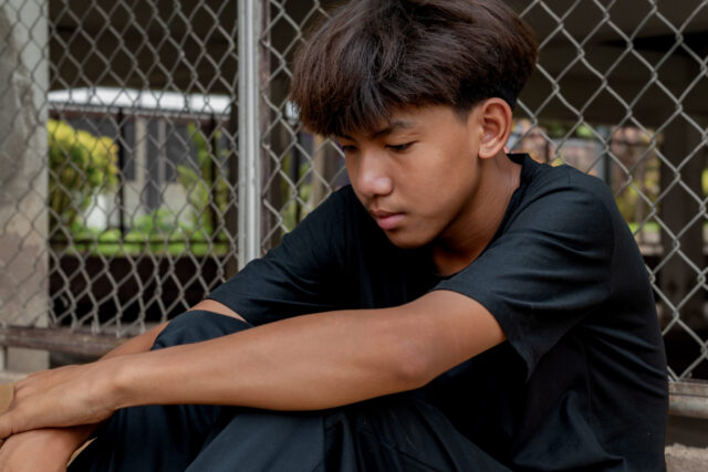 A teenage boy sits with his arms wrapped around his knees, looking down thoughtfully. He has straight brown hair, wears a black t-shirt, and appears worried—perhaps reflecting on a recent visit to juvenile court. A metal fence and building are behind him.