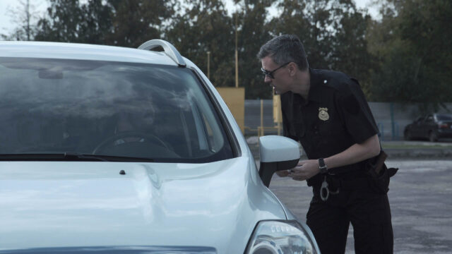 A police officer wearing glasses stands beside a white car and speaks to the driver through the open window during a roadside stop in New York, where reasonable suspicion prompted the interaction amid a tree-lined parking area.