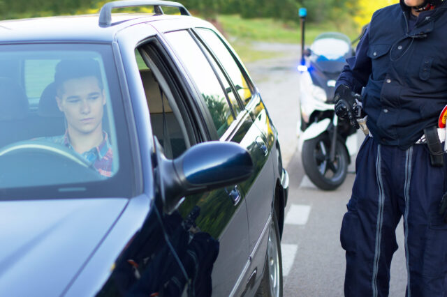 A police officer conducts a search beside a car during a traffic stop in New York, while a young man sits in the driver’s seat looking down. A police motorcycle with flashing lights is parked behind the car on the road.